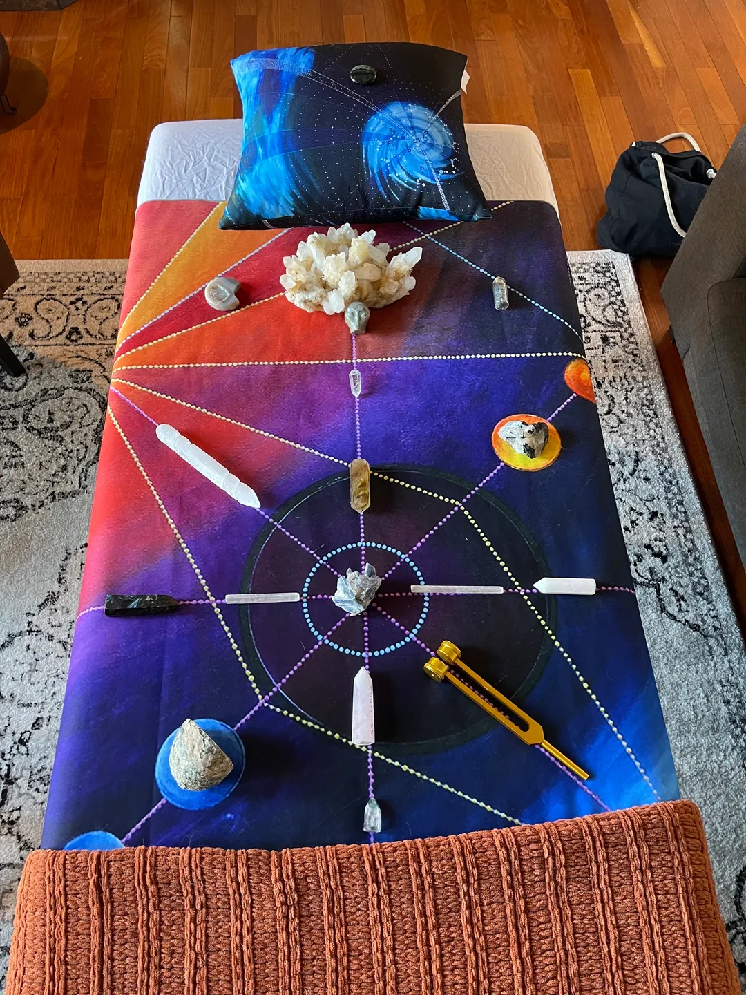 Massage table with a colourful tapestry called Orbital Crown draped over the table, crystals placed in certain grid lines for energy and healing. A colourful pillow with artwork called 