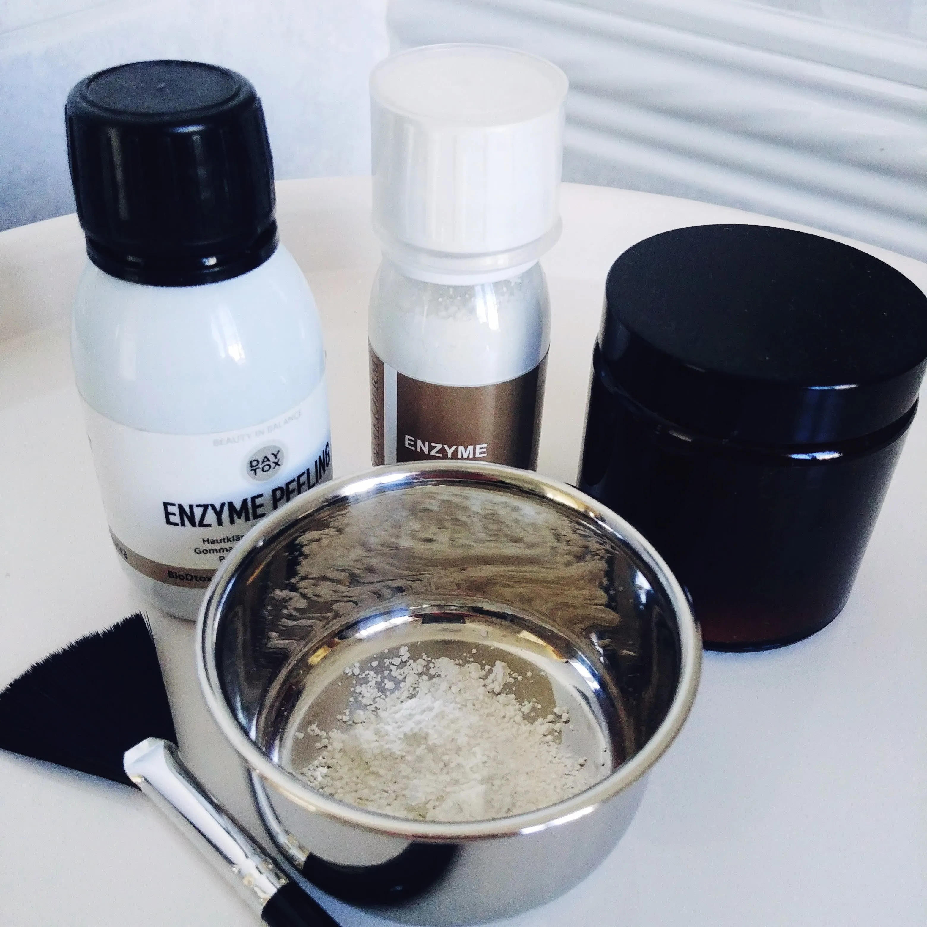 Arrangement aus verschiedenen Enzympeeling Produkten von Daytox, Idealderm mit Pinsel, ein Braunglastigel und einem Schälchen mit Peelingspulver auf weißem Hintergrund. 