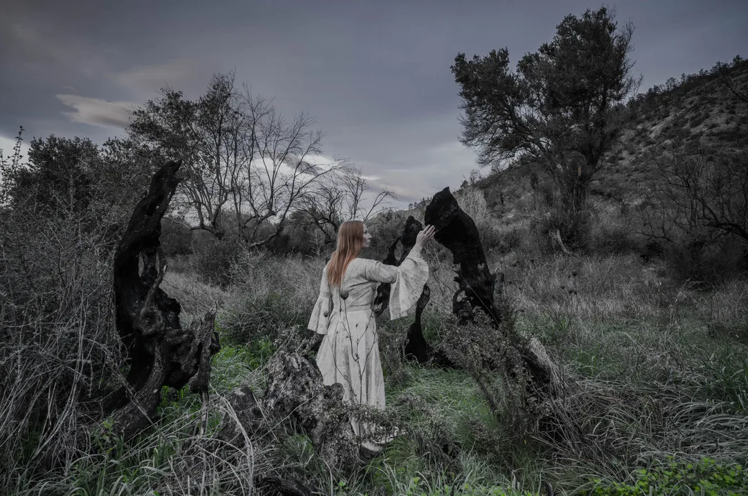 A woman in white stands between two burned tree trunks surrounded by grasses and emerging vegetation. The Bride Tree in Rovies, 2,500 years old