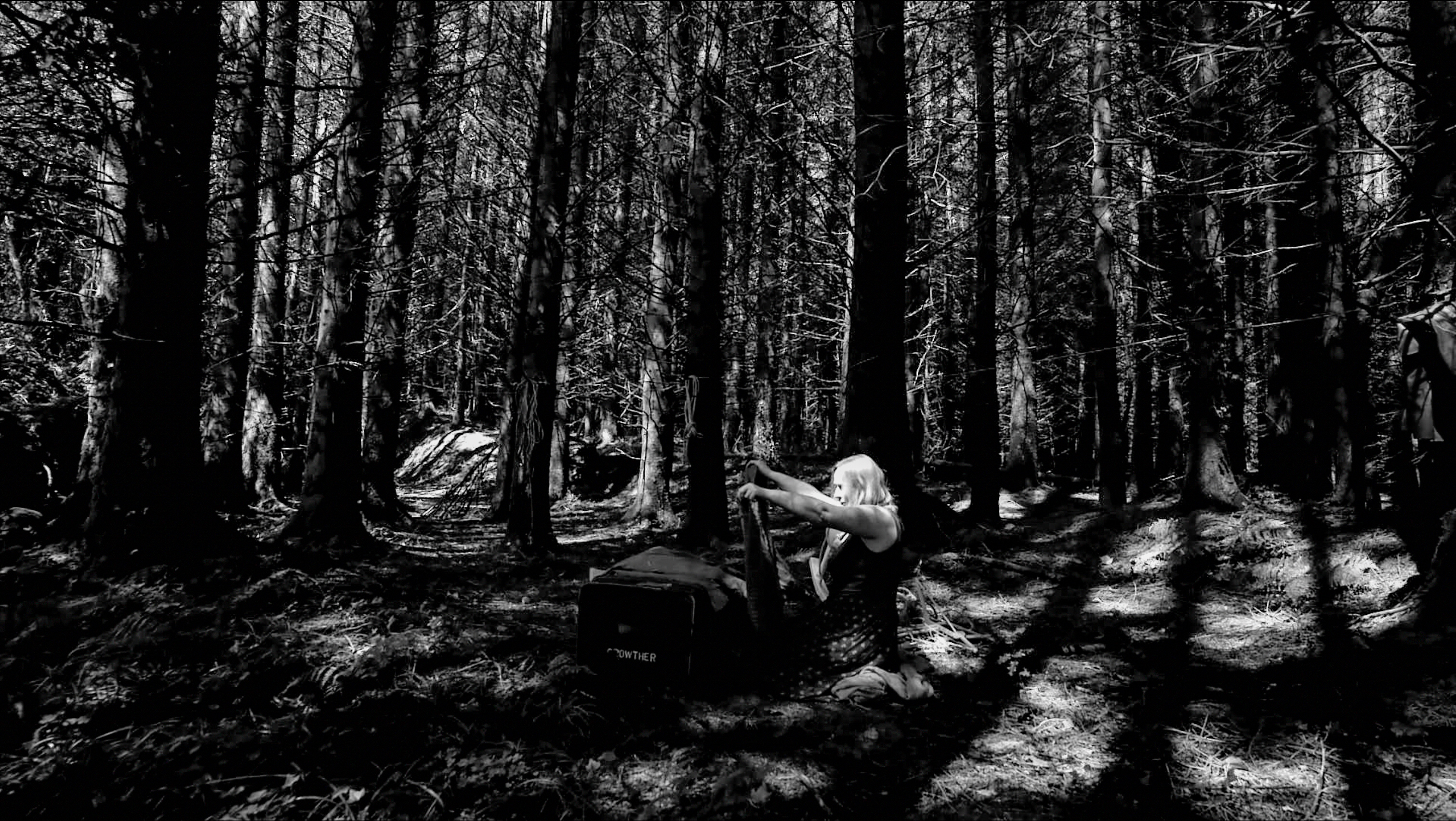Black-and-white image of a woman seated on the forest floor, lifting a garment from an open storage box among tall conifers.
