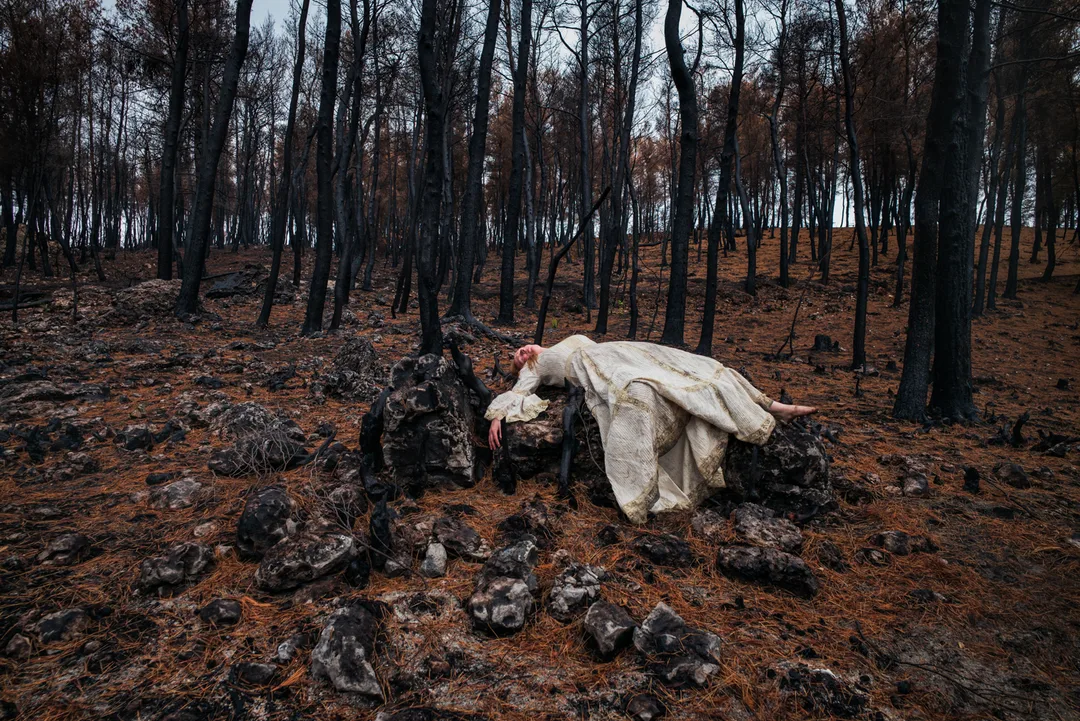 A woman draped in white lies across charred rocks and blackened tree remains in a fire-scarred forest.