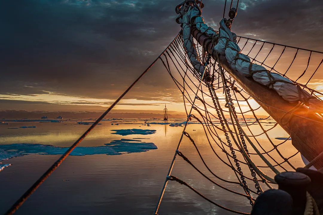 The rigging and netting of a schooner extend over calm water and scattered ice floes as the sun sets across Scoresby Sund.