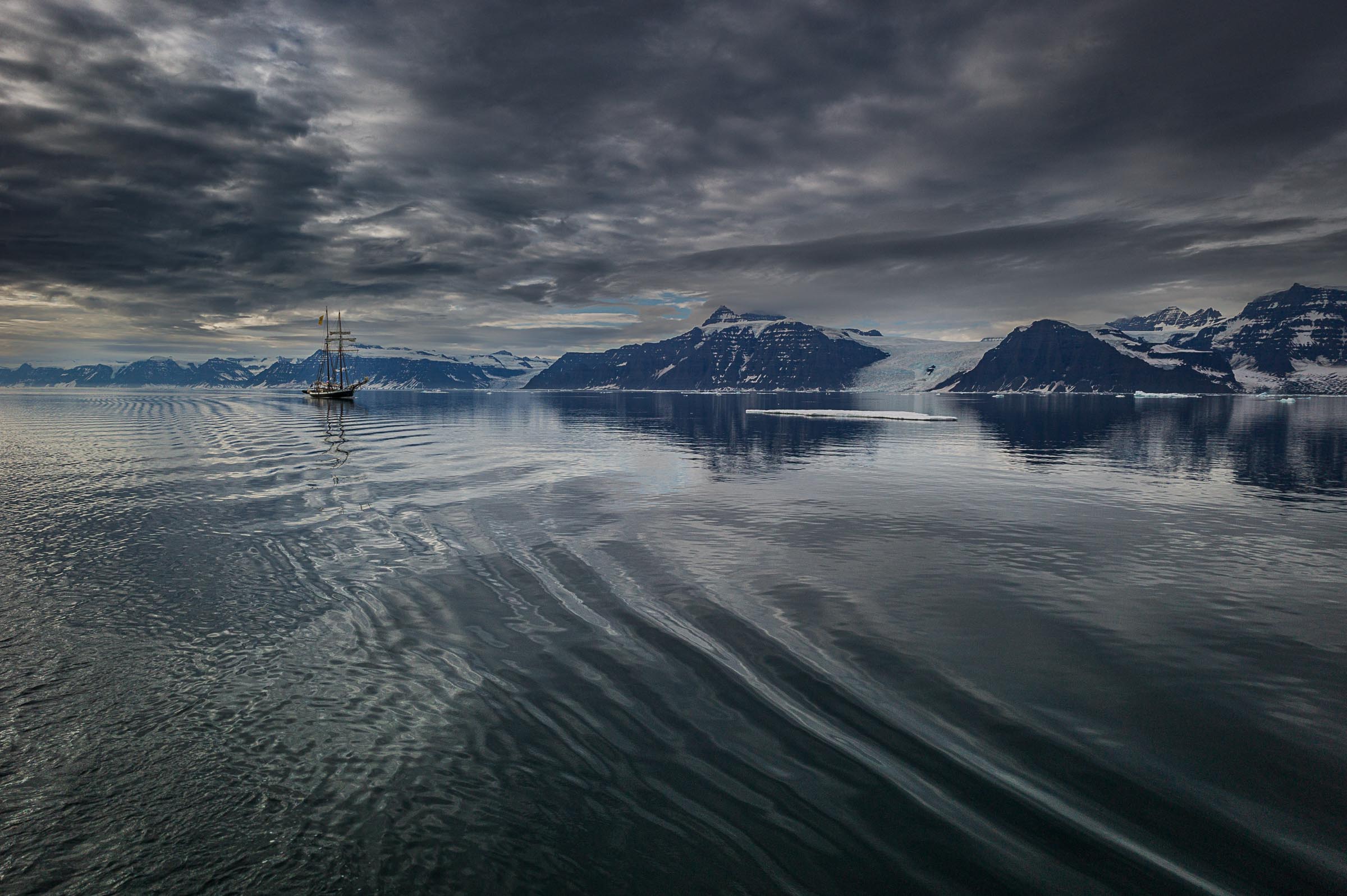 A distant schooner the opal crosses a calm Arctic fjord beneath dramatic layered clouds and snow-covered mountains.
