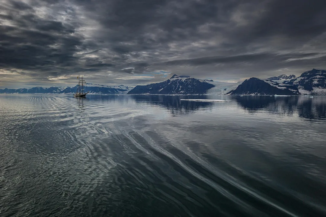 A distant schooner the opal crosses a calm Arctic fjord beneath dramatic layered clouds and snow-covered mountains.
