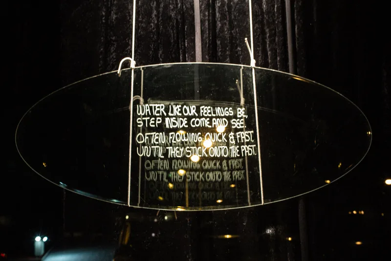 A transparent oval mirror panel carrying handwritten text hangs by cords within a darkened installation space.