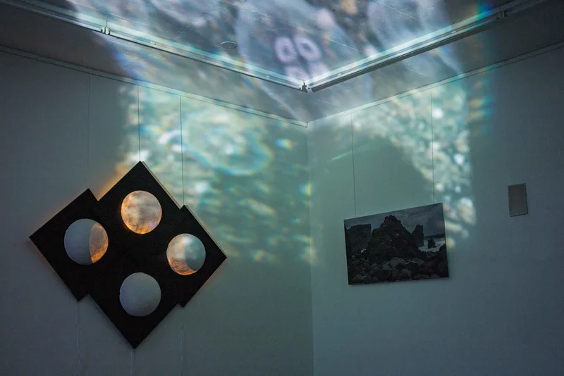 Projected water patterns move across gallery walls and ceiling above a black geometric artwork with circular openings and a smaller dark wall piece.