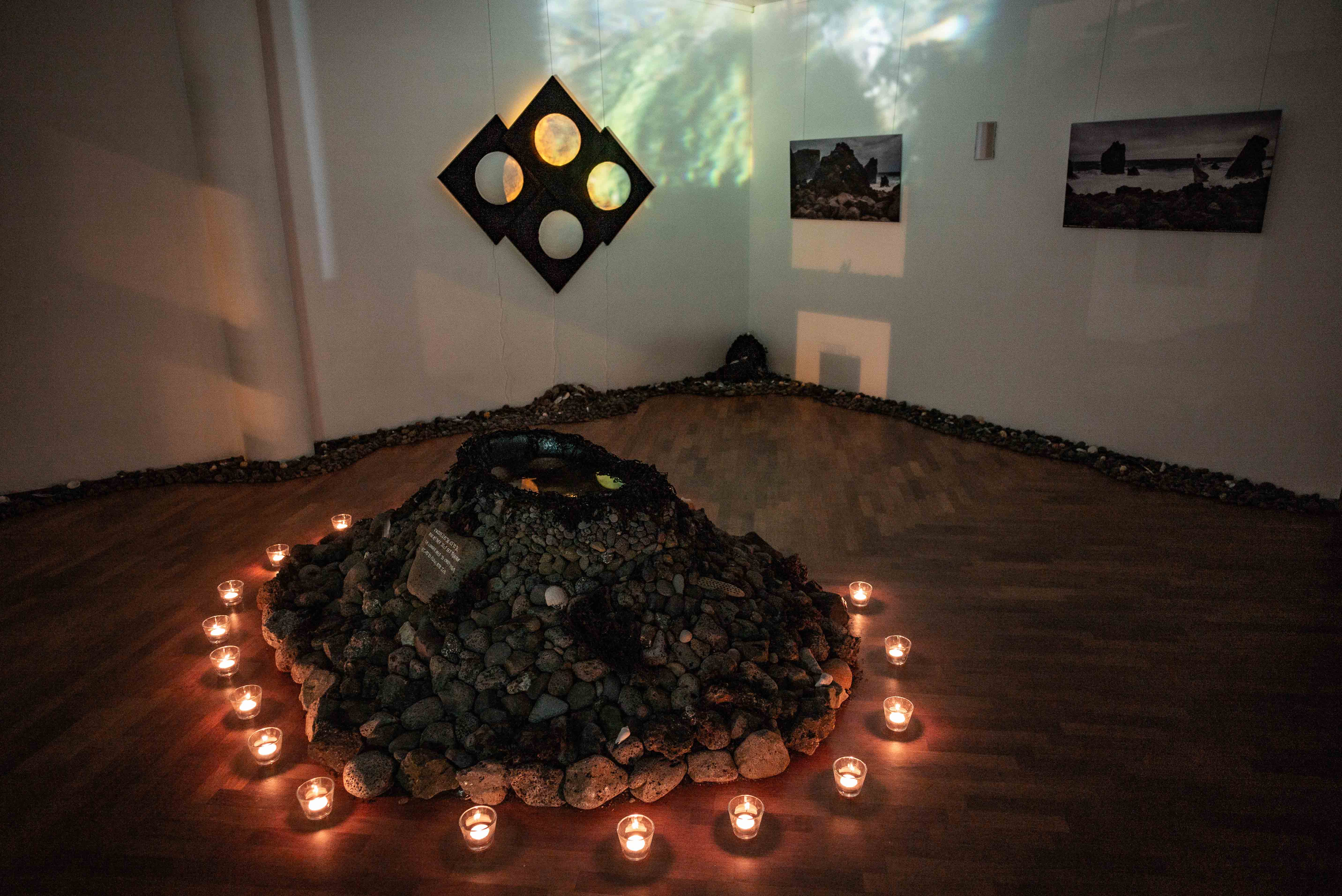 A mound of dark volcanic material is encircled by small glowing candles, with projected imagery and artworks displayed on the surrounding walls.
A flat grey stone bearing handwritten text rests among rounded pebbles and pieces of dried seaweed.