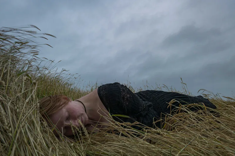 A red-haired woman in a black dress lies curled on her side among tall, dry grass beneath an overcast sky.