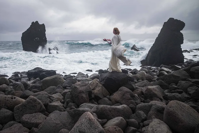 A woman in a flowing white dress walks across a black boulder shoreline between sea stacks as waves break behind her.