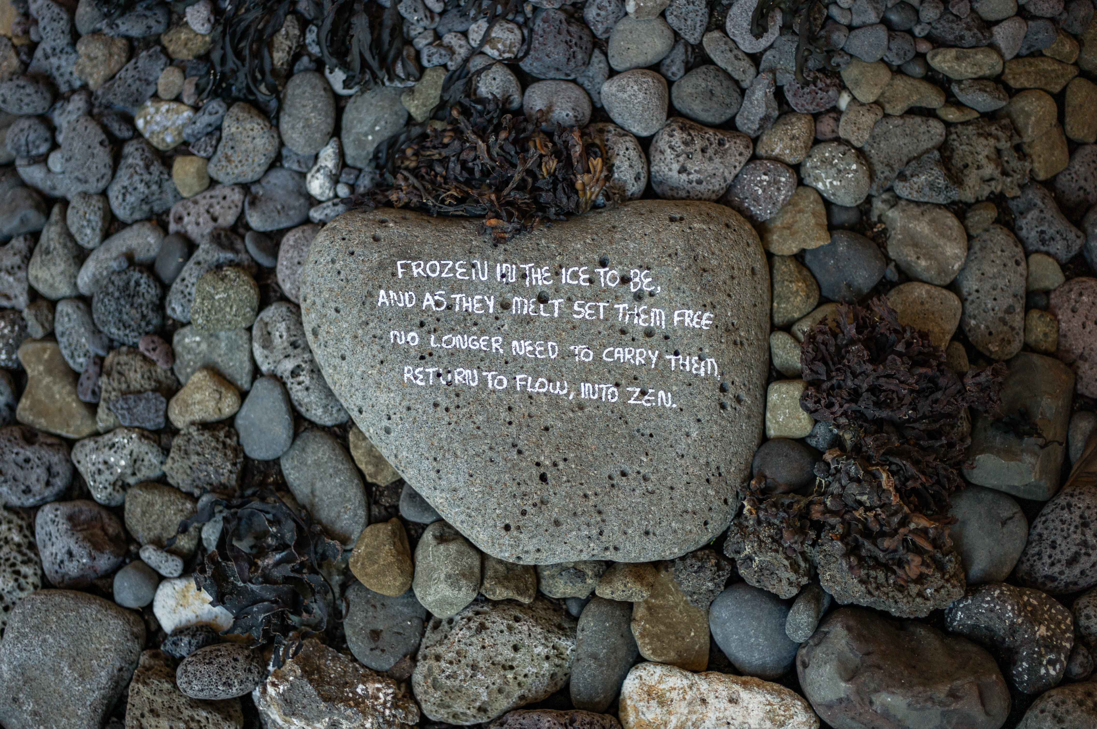 A flat grey stone bearing handwritten text rests among rounded pebbles and pieces of dried seaweed.