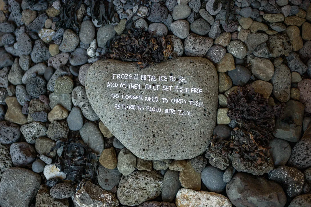 A flat grey stone bearing handwritten text rests among rounded pebbles and pieces of dried seaweed.
