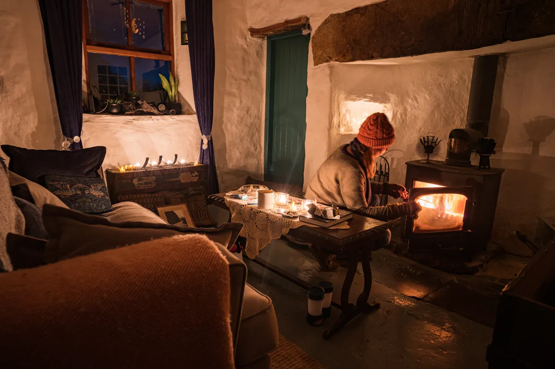 A person sits beside a glowing wood-burning stove inside a warmly lit woodland cottage.