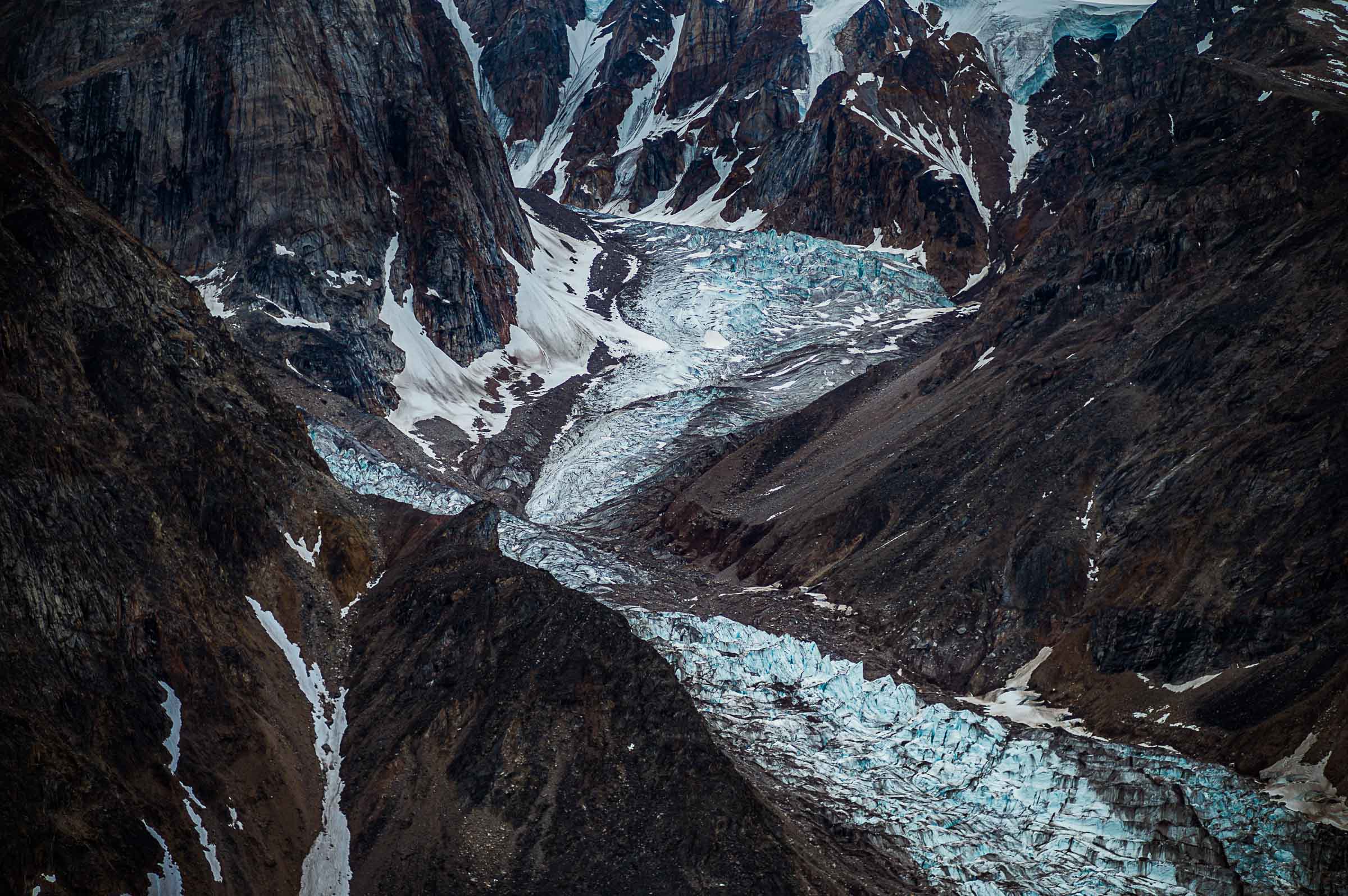 A glacier winds through a steep, dark mountain valley, its fractured blue surface crossed by bands of rock and sediment.

