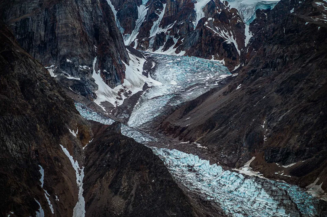 A glacier winds through a steep, dark mountain valley, its fractured blue surface crossed by bands of rock and sediment.
