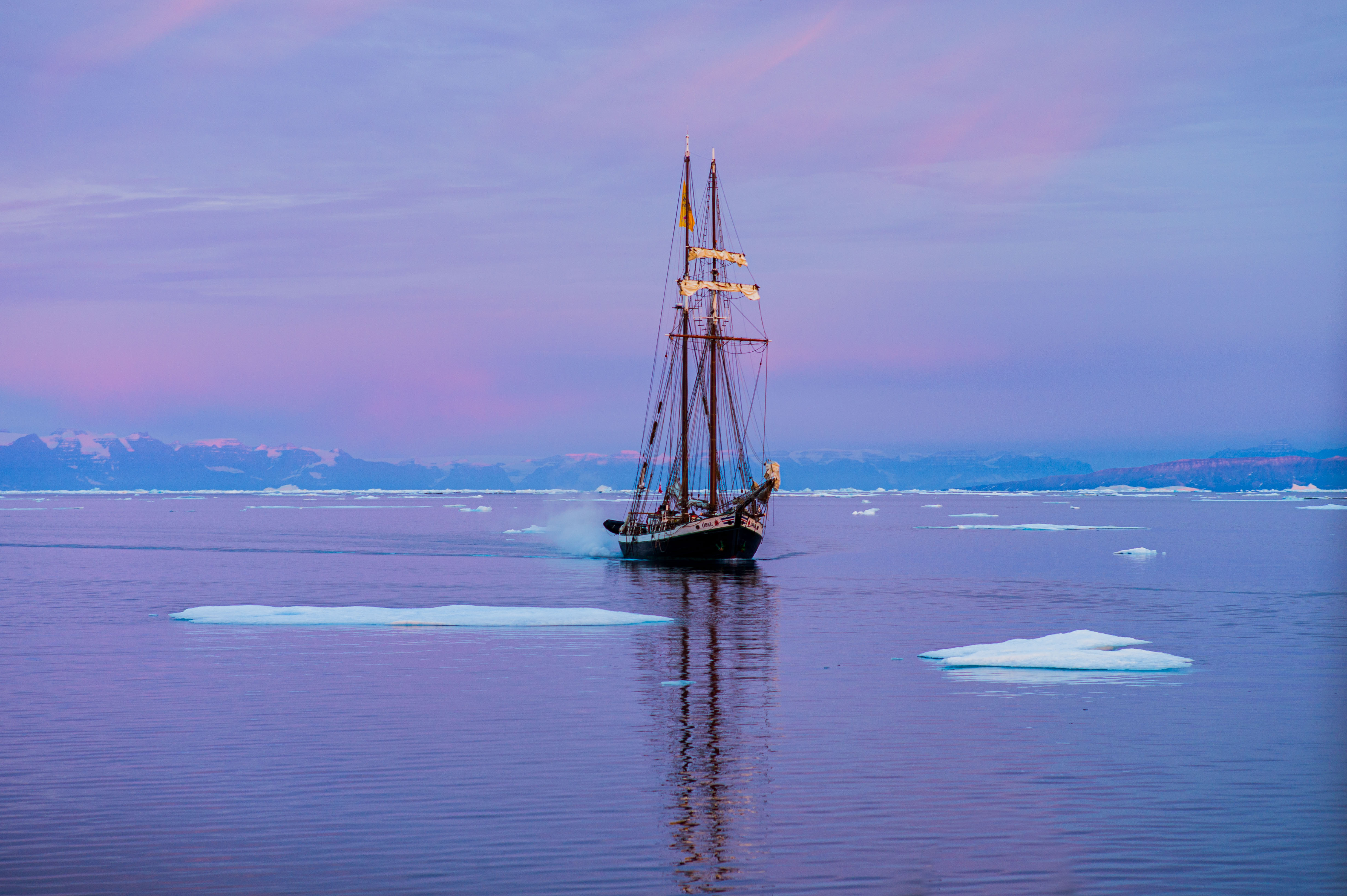 A two-masted schooner among scattered ice floes on calm water beneath a pink and purple Arctic sky. The opal
