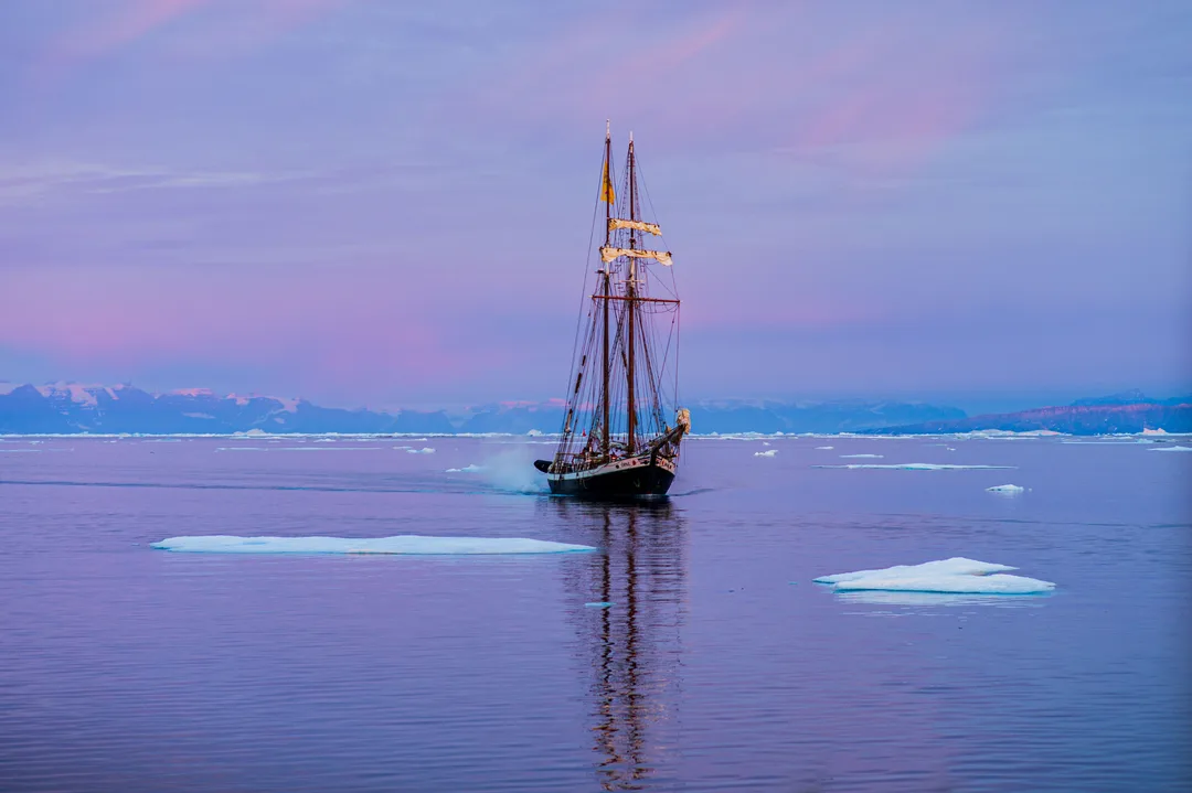 A two-masted schooner among scattered ice floes on calm water beneath a pink and purple Arctic sky. The opal
