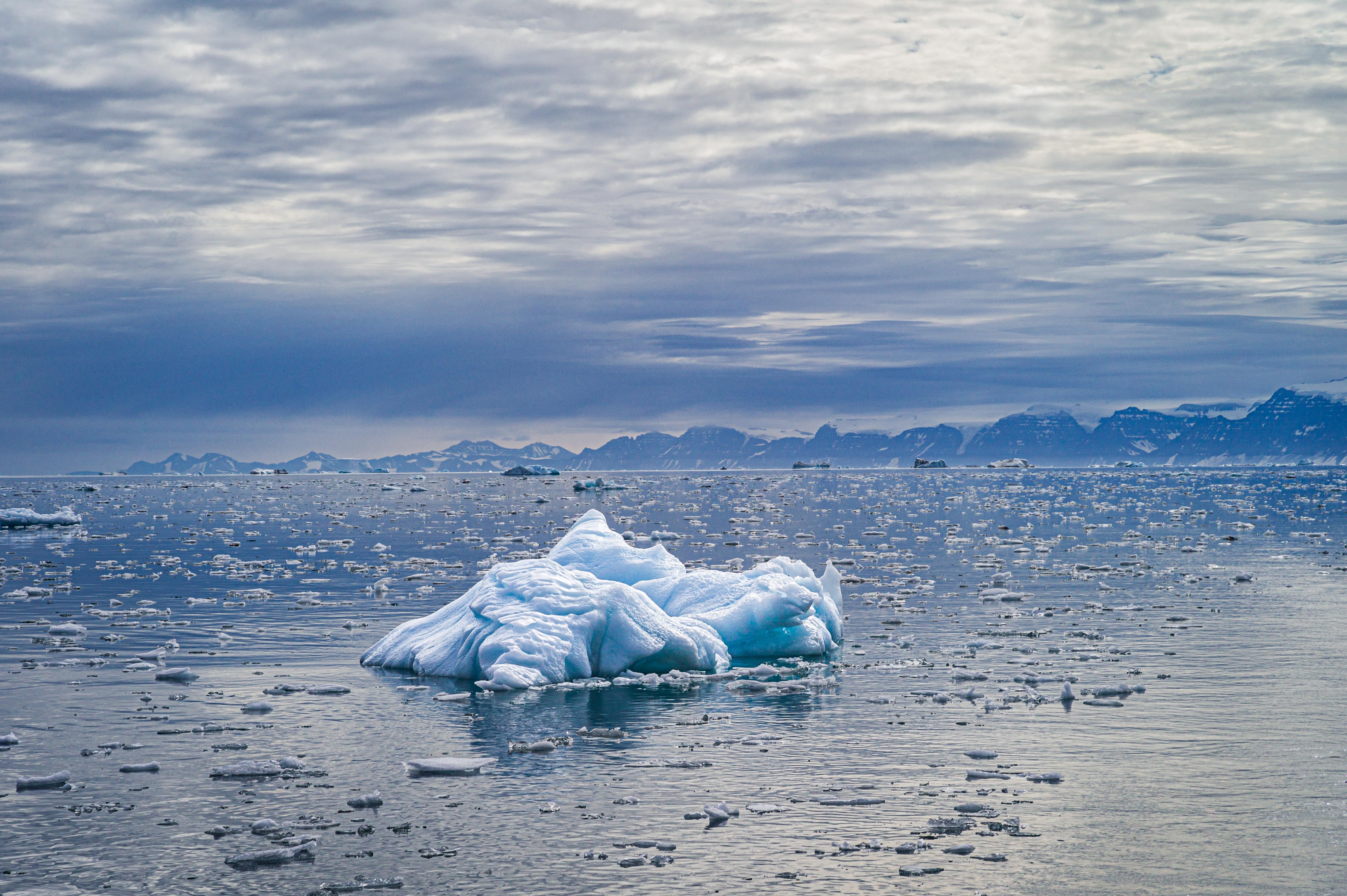 A small sculpted iceberg drifts among countless fragments of sea ice, with distant mountains beneath a layered Arctic sky.
