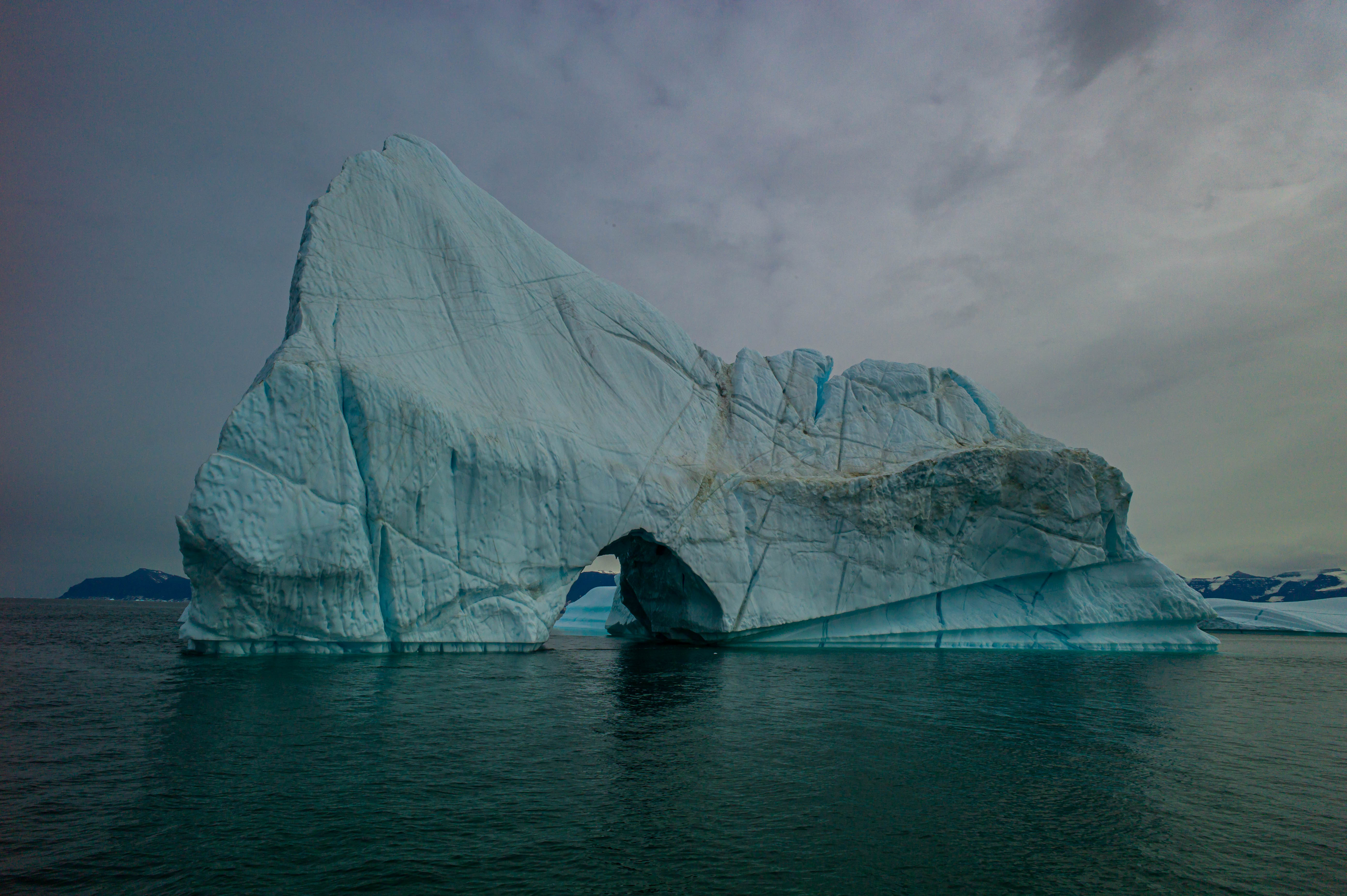 A monumental blue-white iceberg with a large arched opening rises from the dark waters of Scoresby Sund.
