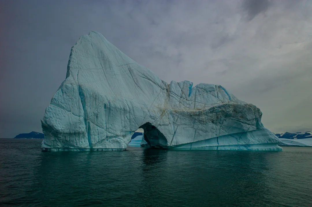 A monumental blue-white iceberg with a large arched opening rises from the dark waters of Scoresby Sund.
