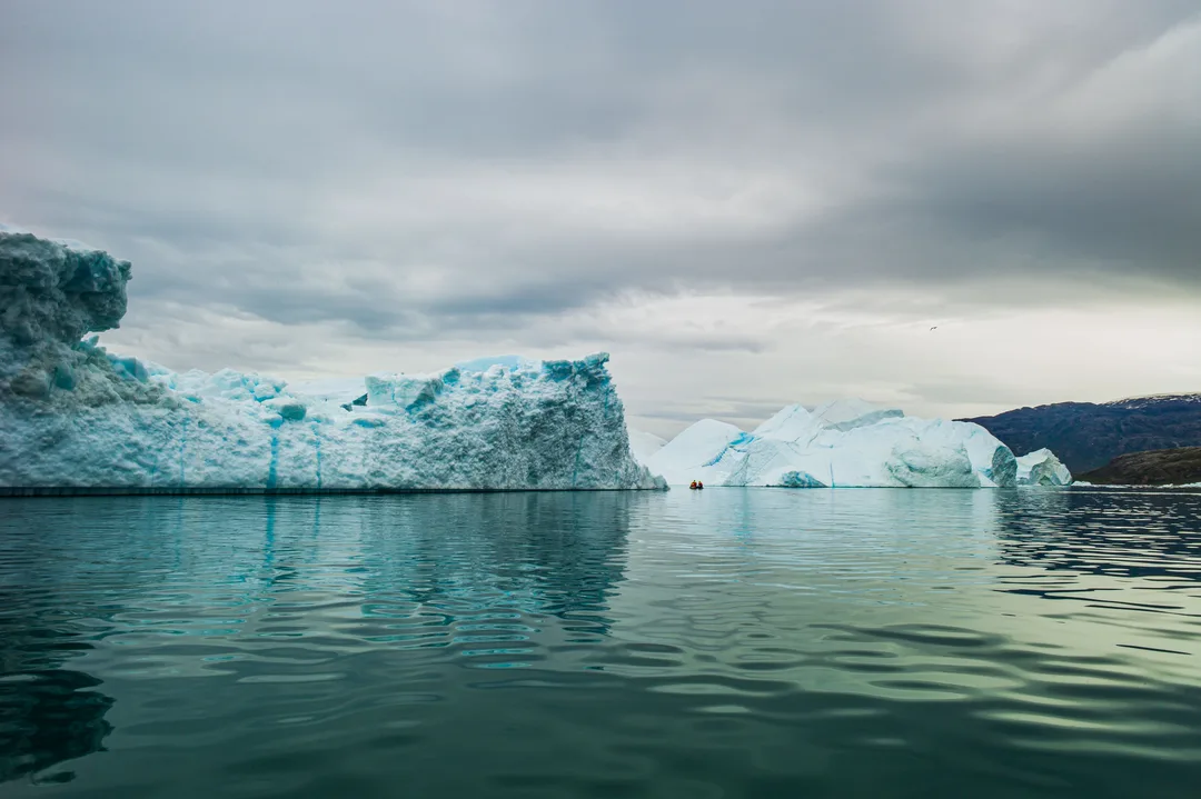 Fractured blue-white icebergs stretch across calm water beneath low grey clouds in Scoresby Sund, East Greenland. A small Zodiac passes between them
