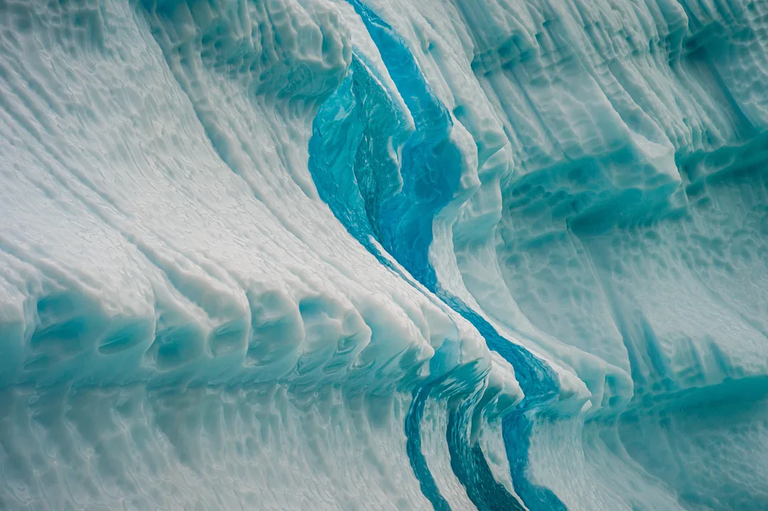 Close view of layered blue and white ice reflected in water, its surface marked by flowing dark lines.
