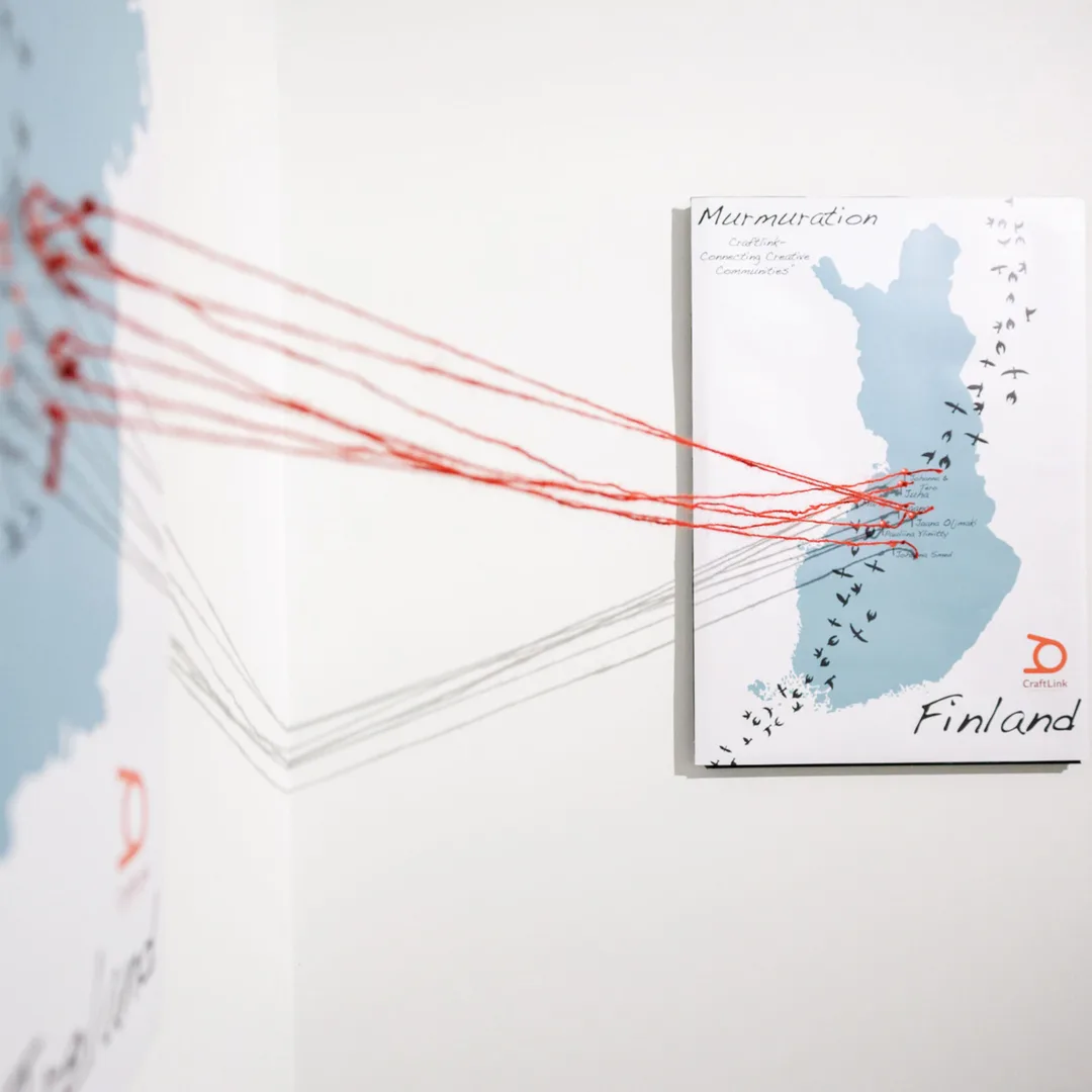 Murmuration installation view showing red threads extending across a white gallery towards a panel mapping the participating Finnish artists.