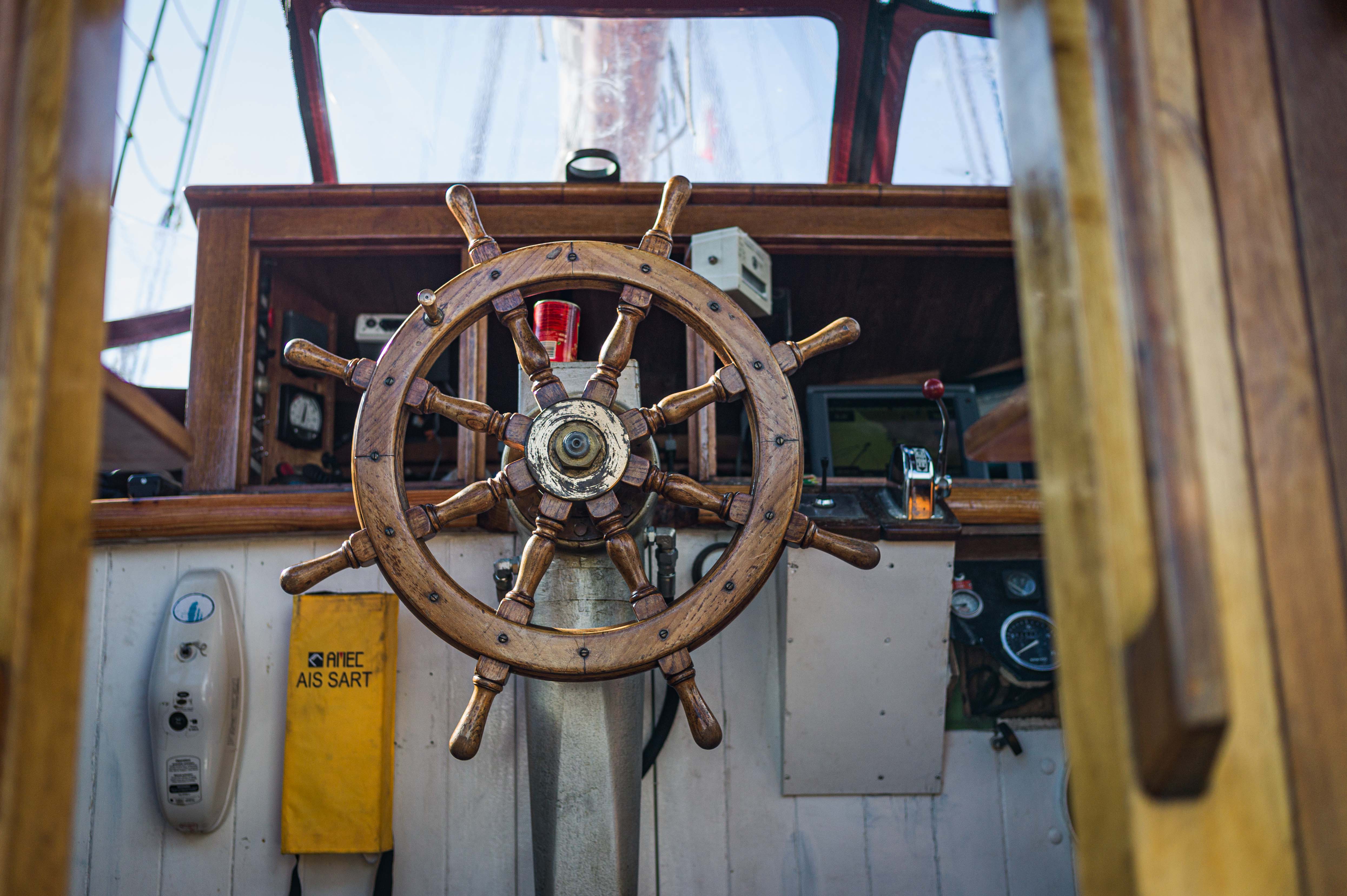 A wooden ship’s wheel at the helm of a sailing vessel, framed by the windows and instruments of the wheelhouse. The Donna Wood North Star Sailing
