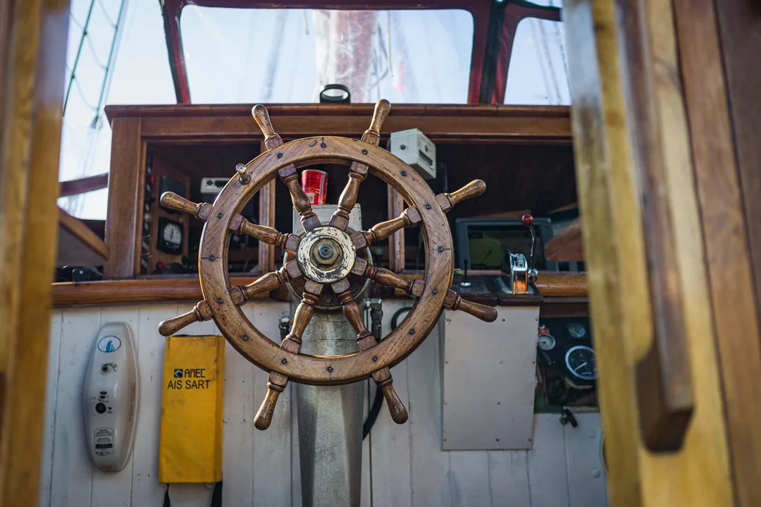 A wooden ship’s wheel at the helm of a sailing vessel, framed by the windows and instruments of the wheelhouse. The Donna Wood North Star Sailing
