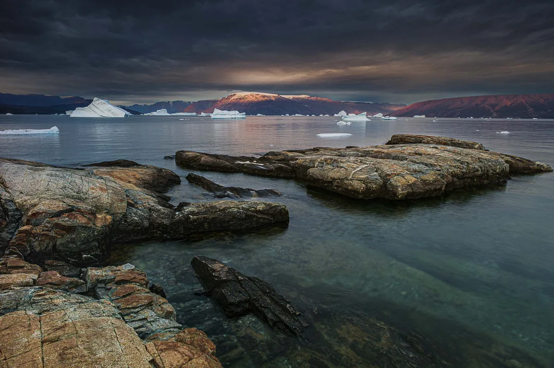 Icebergs drift across a still fjord beyond weathered shoreline rocks as warm evening light breaks beneath dark storm clouds.
