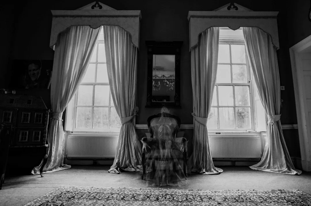 Black-and-white still from Trinity, showing Aishling Muller seated between two curtained windows, with layered figures appearing within the room.