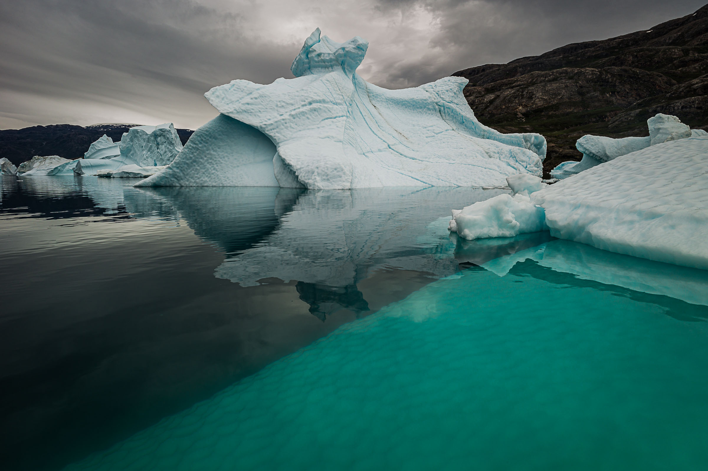 A sculpted iceberg beside vivid turquoise water and a dark rocky shoreline in Scoresby Sund.
