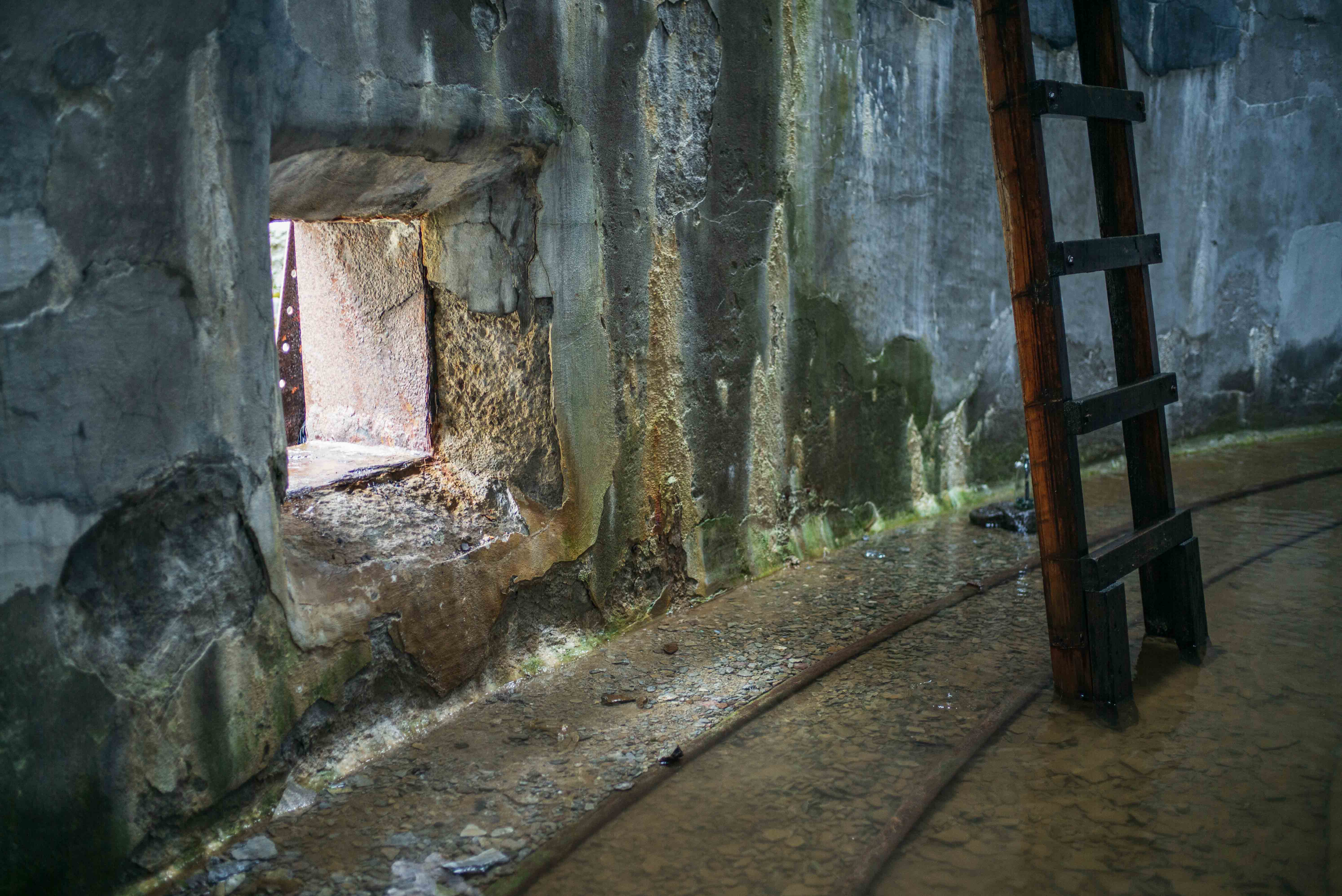 View through a low opening inside a corroded storage tank towards the damaged factory interior, with a dark ladder standing to the right.