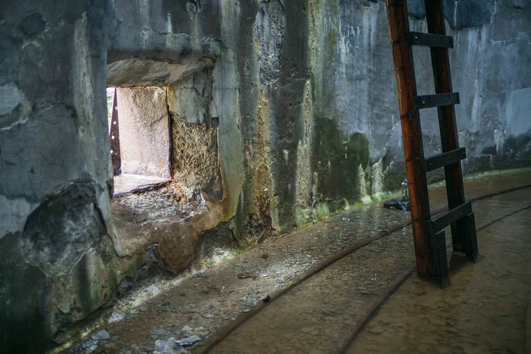 View through a low opening inside a corroded storage tank towards the damaged factory interior, with a dark ladder standing to the right.