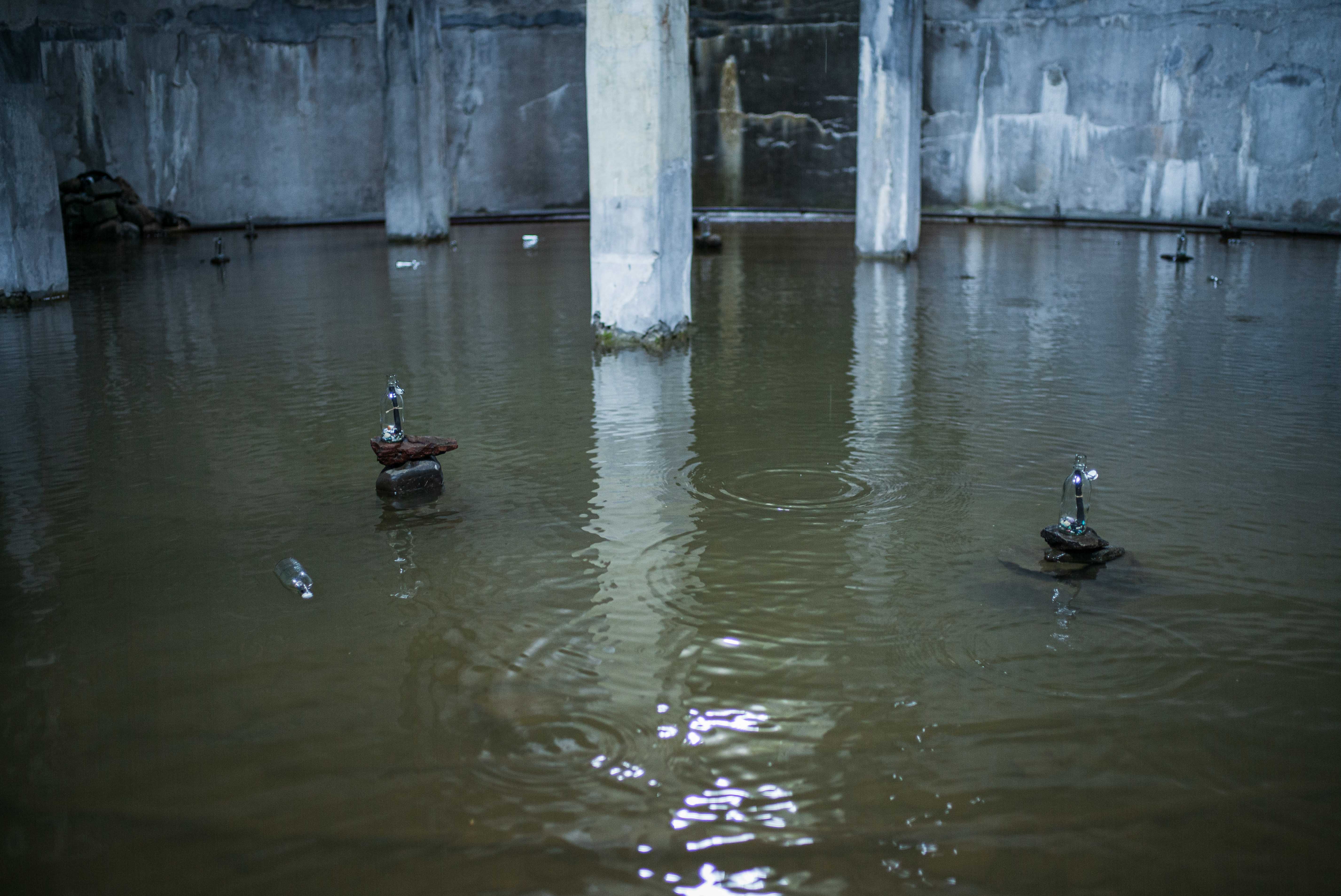 Three small sculptures made from bottles, stones and found materials stand in shallow water across the flooded factory floor.