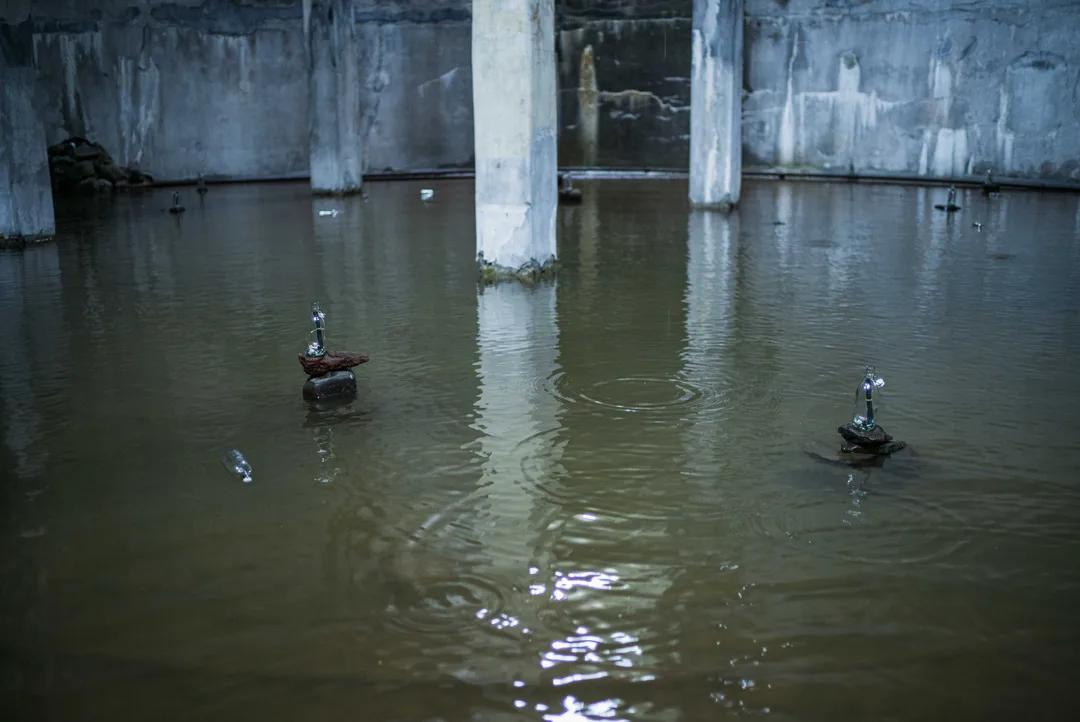 Three small sculptures made from bottles, stones and found materials stand in shallow water across the flooded factory floor.