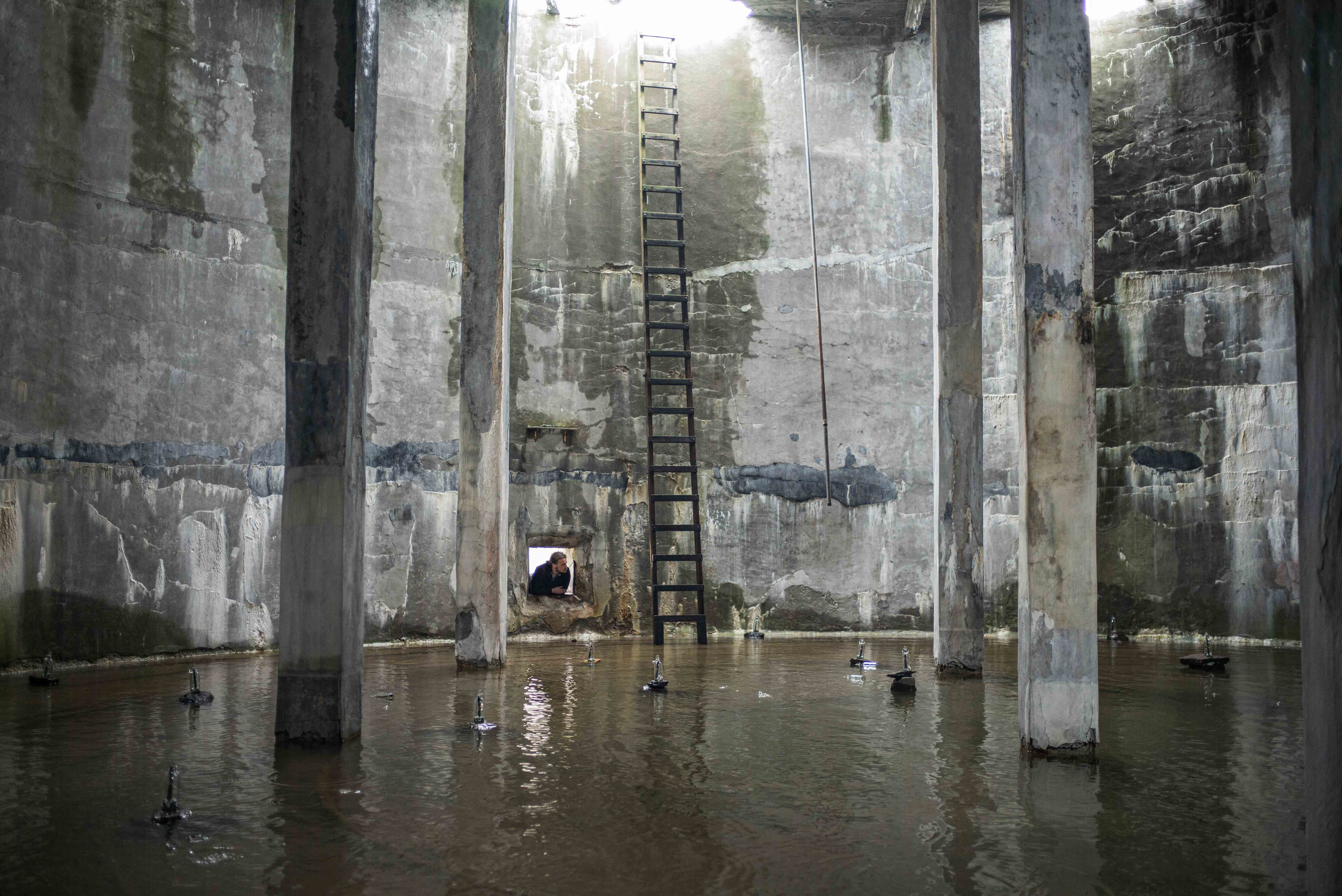 Flooded interior of an abandoned herring factory, with concrete pillars, a rusted storage tank and small sculptural elements reflected in the water.