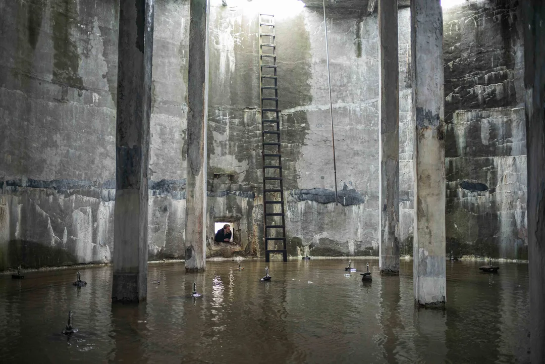 Flooded interior of an abandoned herring factory, with concrete pillars, a rusted storage tank and small sculptural elements reflected in the water.