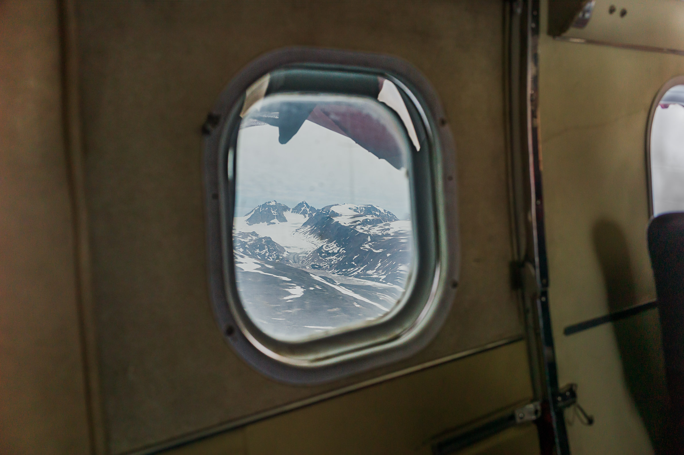 Snow-covered mountains seen through the rounded window of a small aircraft flying over East Greenland.