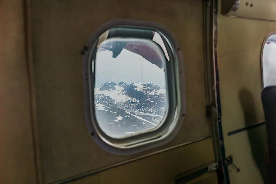Snow-covered mountains seen through the rounded window of a small aircraft flying over East Greenland.