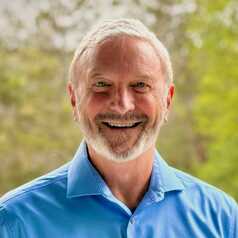 Dr. Paul Thomas, board-certified pediatrician and co-founder of Kids First 4Ever, smiling in a blue collared shirt against a soft green outdoor background.
