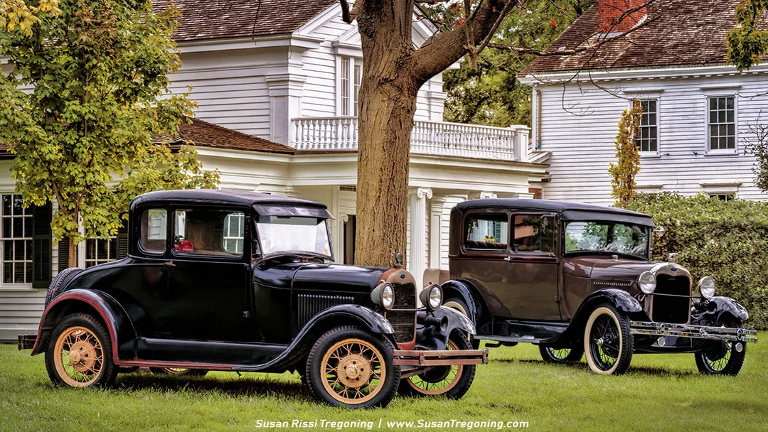 Two Ford Model A automobiles—a dark coupe and a lighter‑colored Tudor sedan—are parked side by side on a grassy lawn beneath a large shade tree. Both cars feature the rounded fenders, spoked wheels, and upright profiles typical of late‑1920s and early‑1930s styling. Behind them stand two white colonial‑style houses with dark shutters and traditional porches, creating a nostalgic historic‑village atmosphere.