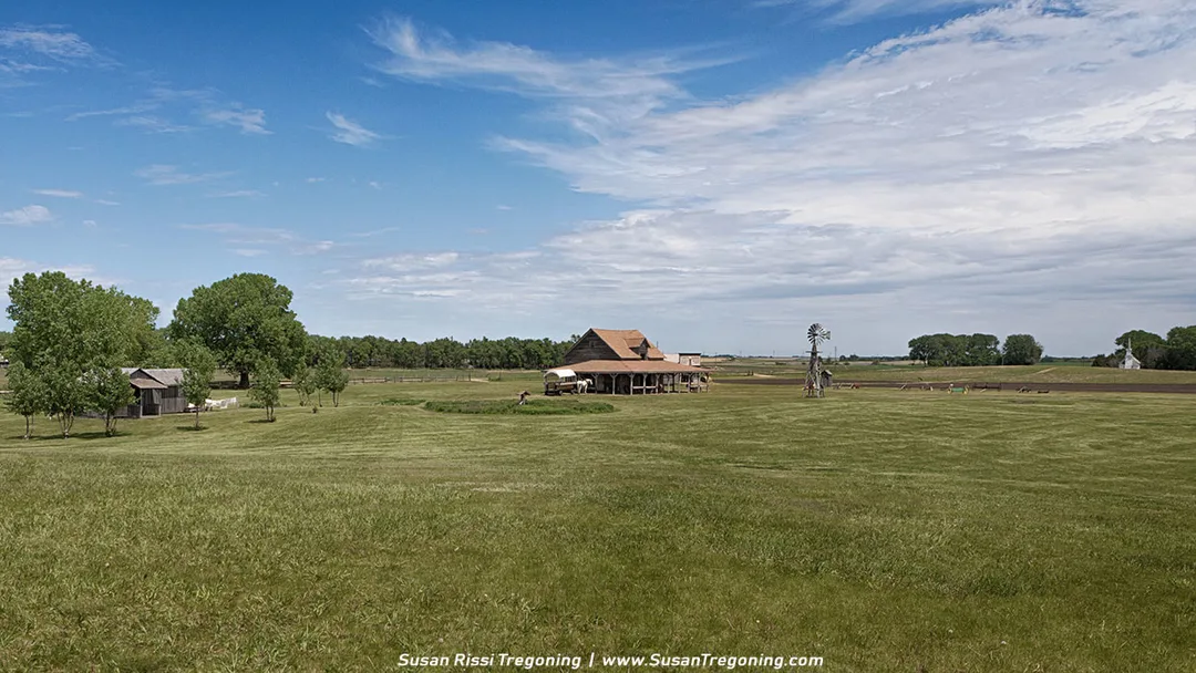 A rural homestead scene showing a large barn, a modest claim shanty, and a windmill surrounded by broad stretches of green prairie.