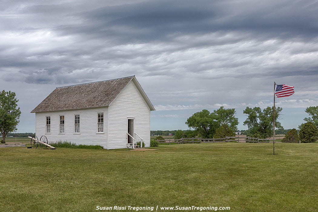 The one‑room schoolhouse on the Charles Ingalls Homestead, now home to the Laura Ingalls Wilder Exhibit, set beside a teeter‑totter and an American flag on the open prairie.