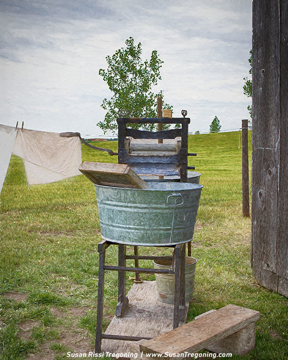 A galvanized metal wash tub on a stand with a hand‑cranked wringer and a wooden washboard, set in a grassy yard with white laundry hanging on a clothesline behind it.