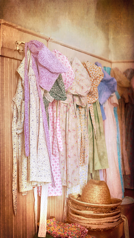 A collection of small calico dresses and sunbonnets hanging in a schoolhouse interior, evoking the clothing worn by Laura Ingalls–era prairie girls.