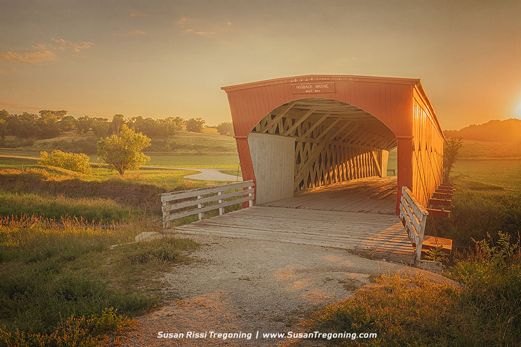 A red covered bridge marked “Hogback Bridge, Built 1884” stands over a rural road at sunset, its white railings leading into a wooden lattice interior. Golden light washes across the surrounding green fields and trees, creating a warm, peaceful evening scene.