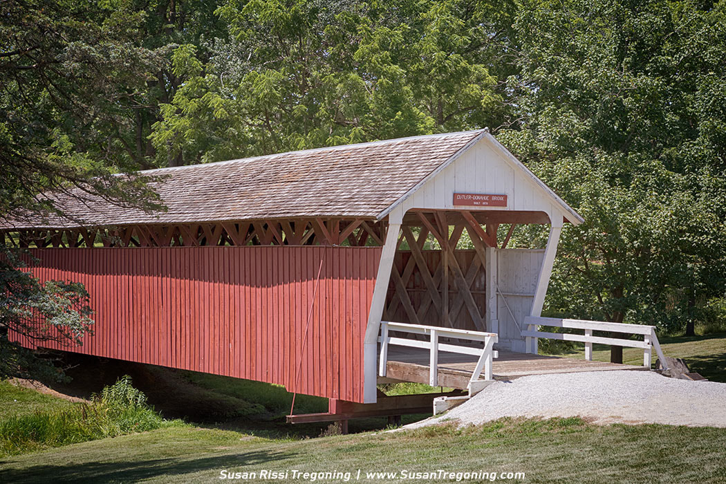 A red covered bridge labeled “Cutler-Donahoe Covered Bridge, Built 1883” spans a small stream in a lush wooded setting, with white trim, a shingled roof, and lattice trusses visible through the side openings. A gravel path leads to the entrance, surrounded by dense green trees and vegetation.