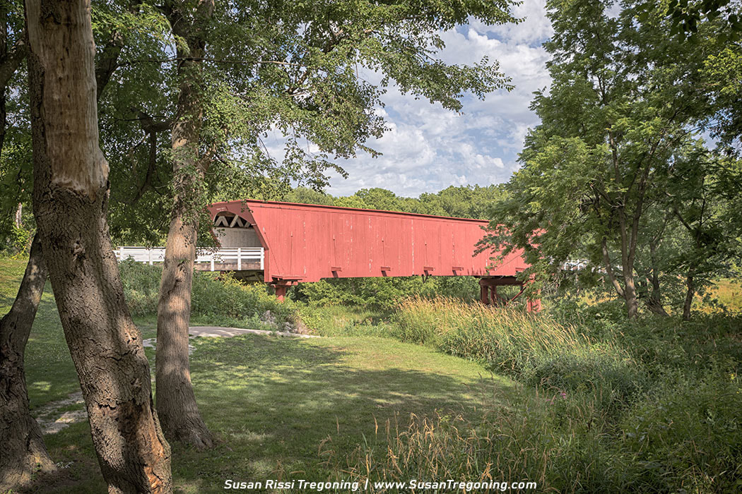 A bright red covered bridge spans a narrow creek in a lush green landscape. The wooden bridge has a pitched roof, white trim at the entrance, and sits on timber supports above the water. Trees with early‑season foliage surround the scene under a partly cloudy sky. 