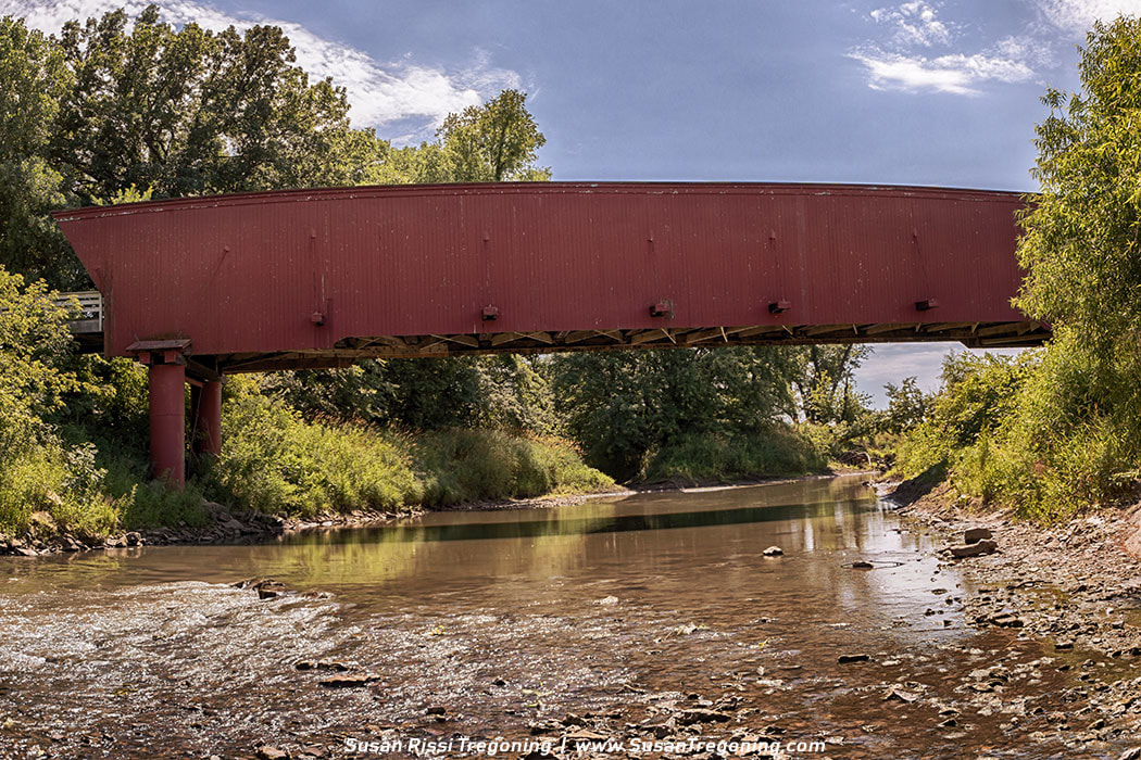 The 1880 Holliwell Covered Bridge stands in a quiet bend of the Middle River, its red siding and timber trusses anchoring the rural Iowa landscape.
