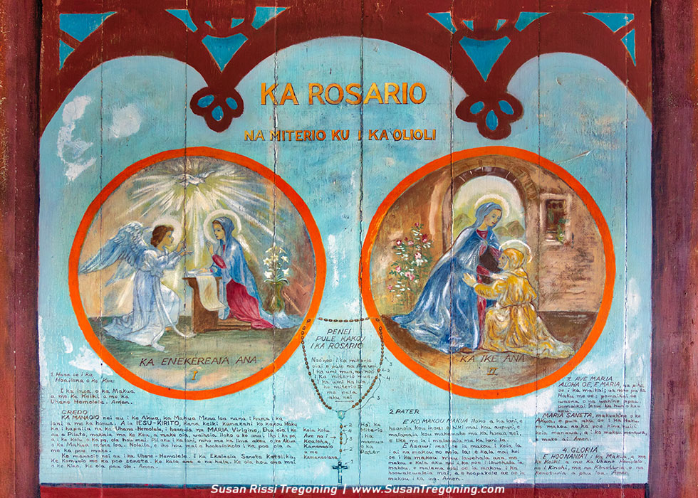 A painted mural titled “Ka Rosario” features two circular scenes from Christian tradition. The left circle shows the Annunciation, with the angel Gabriel appearing to Mary, and the right circle shows the Visitation, with Mary greeting Elizabeth. Hawaiian text surrounds the images, including prayers and instructions for reciting the rosary. Decorative borders and lettering frame the artwork, which blends Catholic iconography with Hawaiian language.